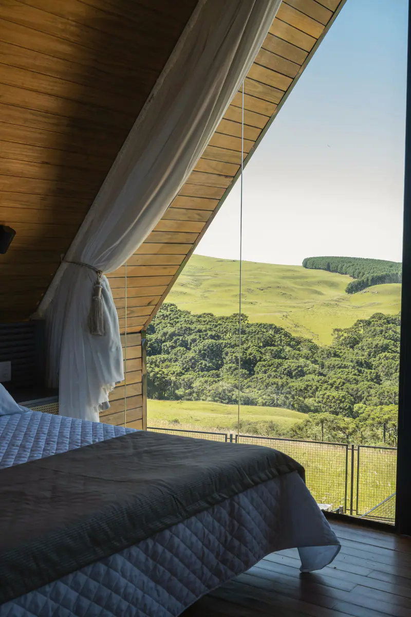 Quarto do Chalé Mirante com parede de vidro e vista para morros verdes e araucárias